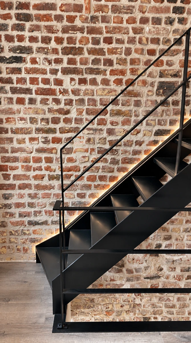 Zwart stalen trap met ledverlichting langs de wang, voor een muur van oude bakstenen